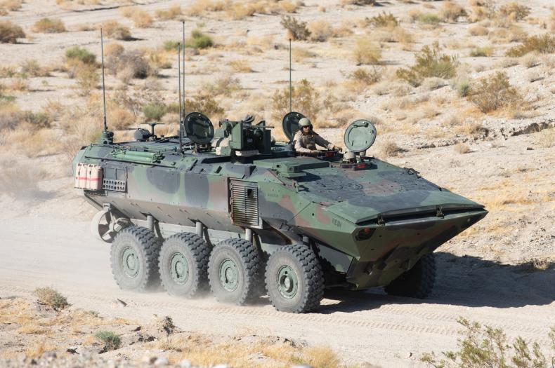 An armored military vehicle with eight wheels speeds through a desert landscape. It features various mounted equipment, and a soldier in protective gear is visible in the gunner's station.